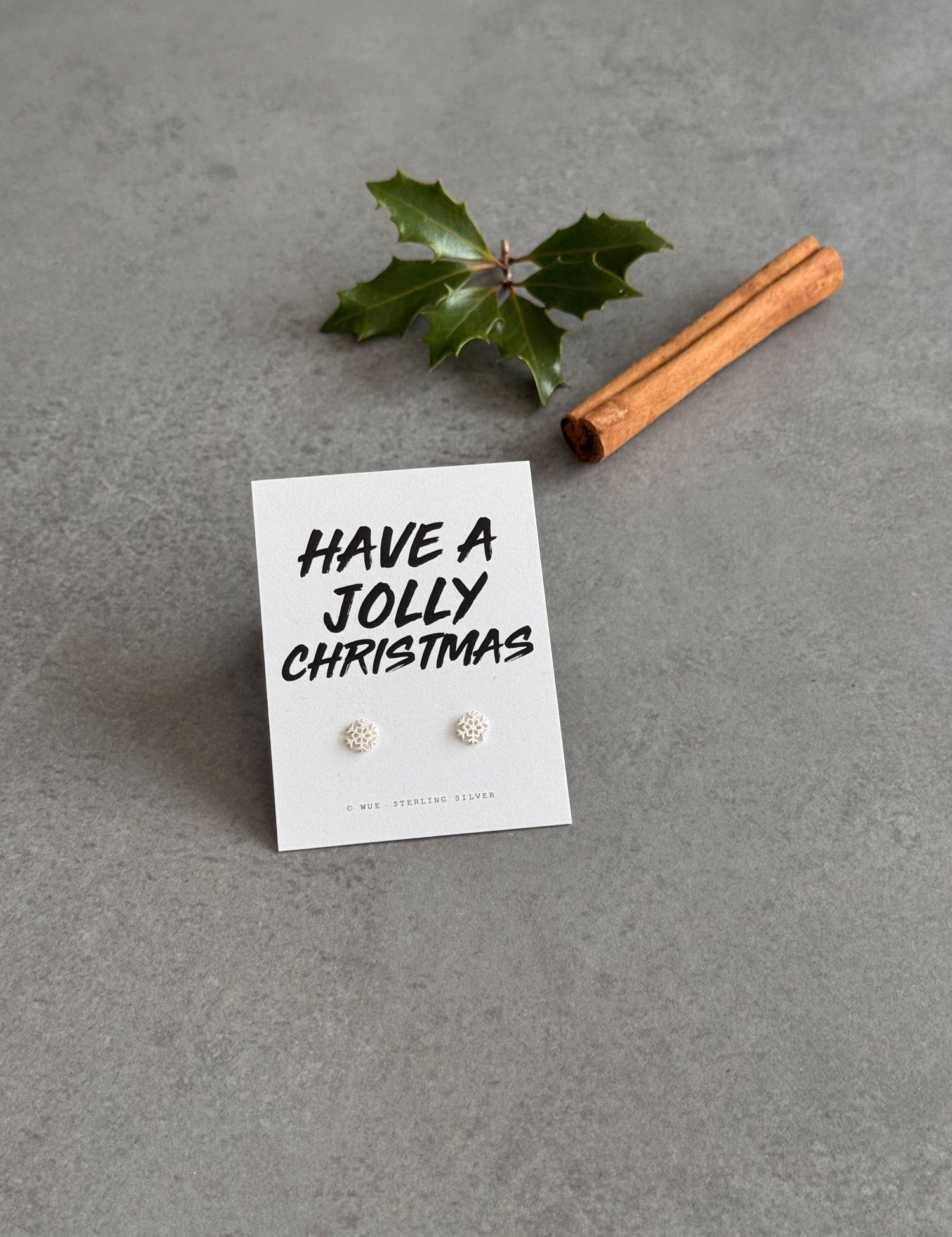 Earring set on a card with 'Have a Jolly Christmas' text, accompanied by a cinnamon stick and holly leaf on a gray background.