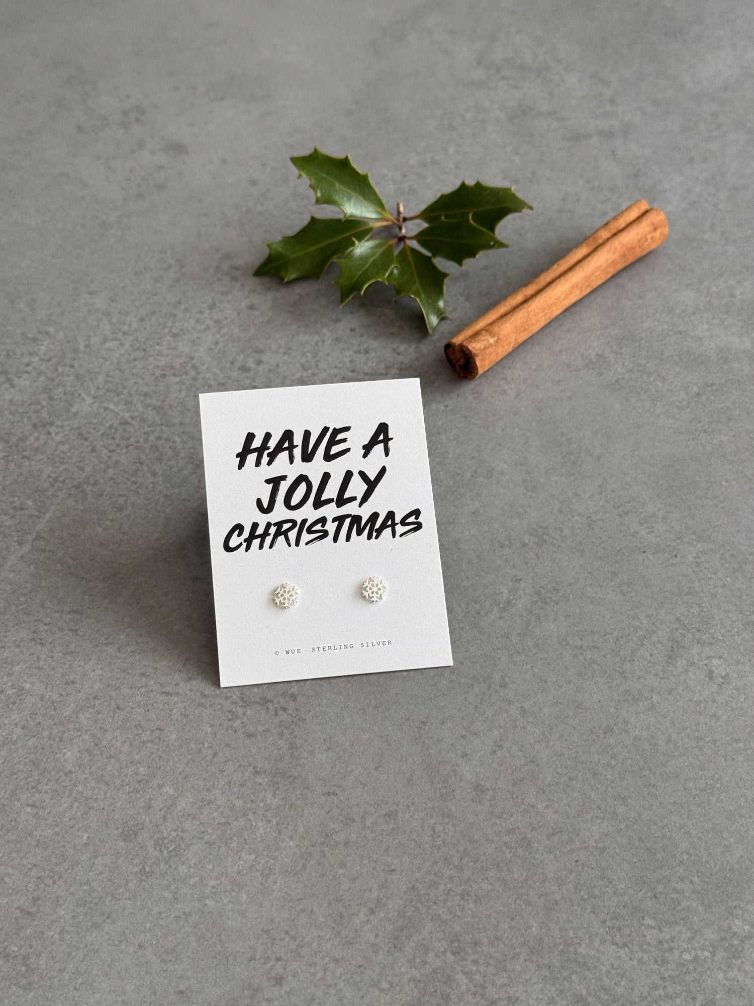 Earring set on a card with 'Have a Jolly Christmas' text, accompanied by a cinnamon stick and holly leaf on a gray background.