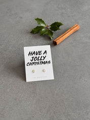 Earring set on a card with 'Have a Jolly Christmas' text, accompanied by a cinnamon stick and holly leaf on a gray background.