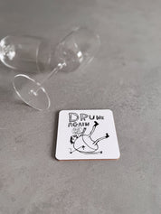 Coaster with 'Drunk Again' text and illustration on a gray surface with a glass.
