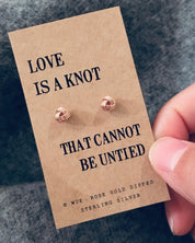 Rose gold earrings on a 'Love is a knot' card held by a hand against a gray background