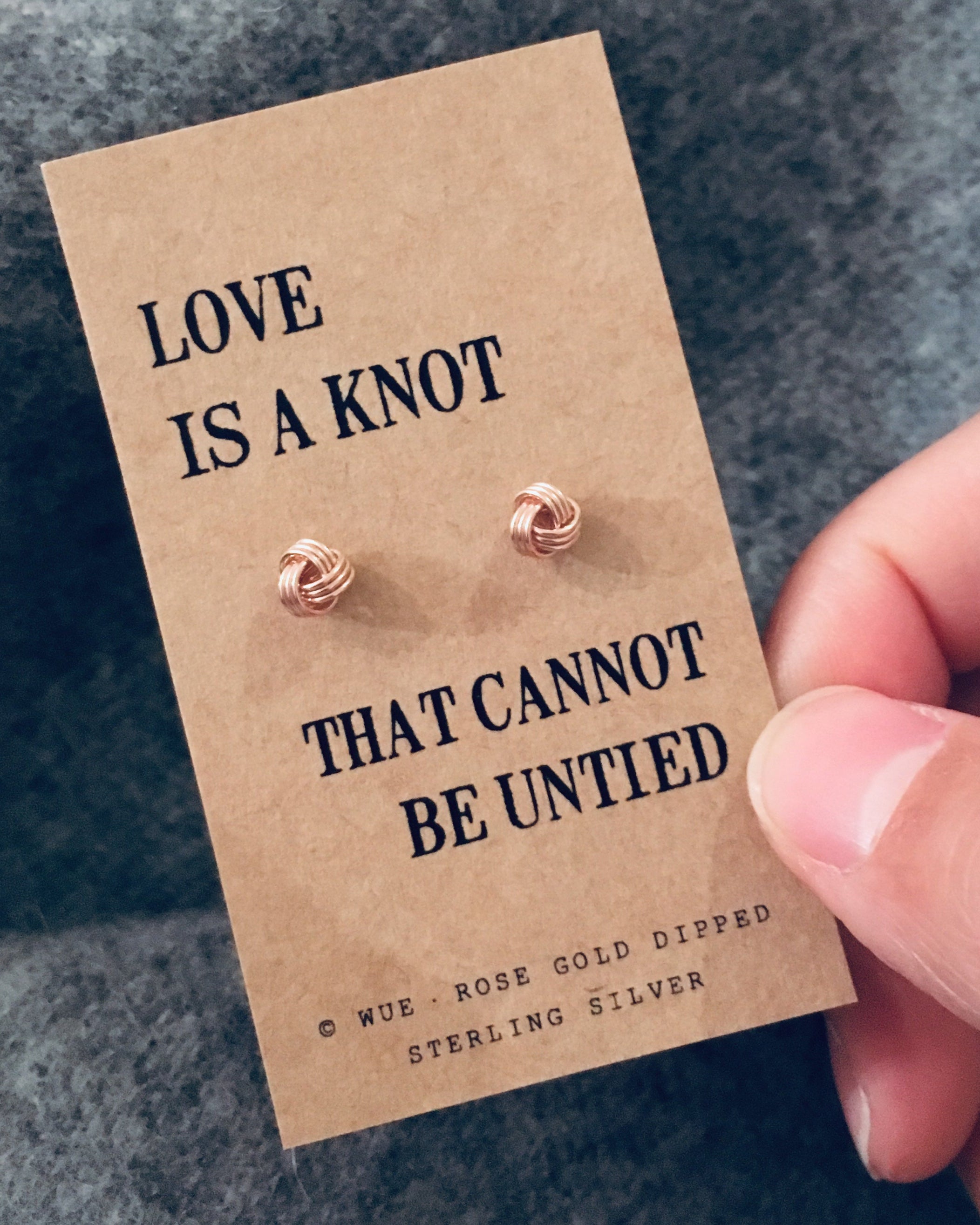 Rose gold earrings on a 'Love is a knot' card held by a hand against a gray background